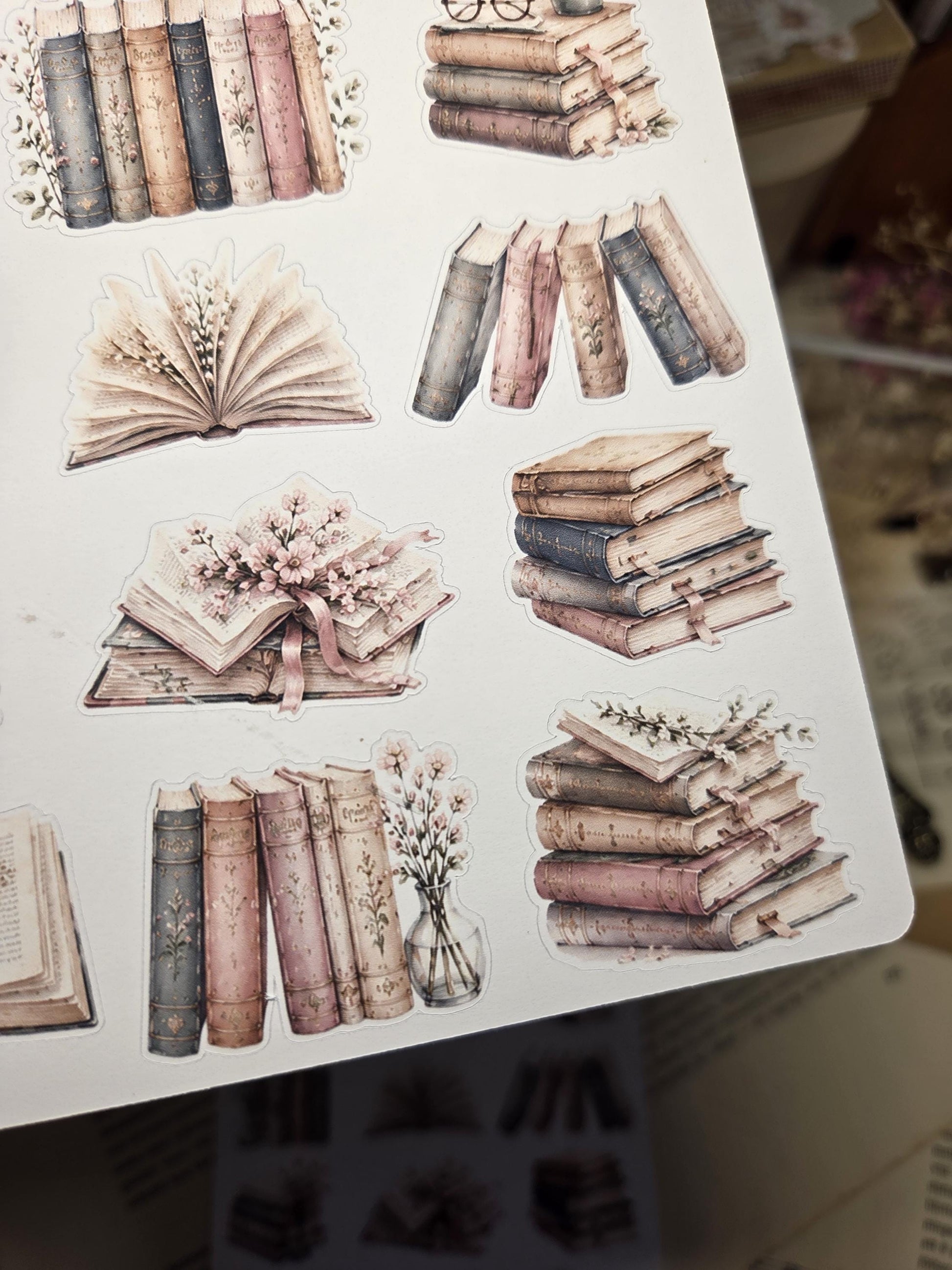 A sticker sheet with illustrations of antique books. The images show stacks of books and individual volumes, decorated with flowers and soft colours. Some books are open, revealing their pages.